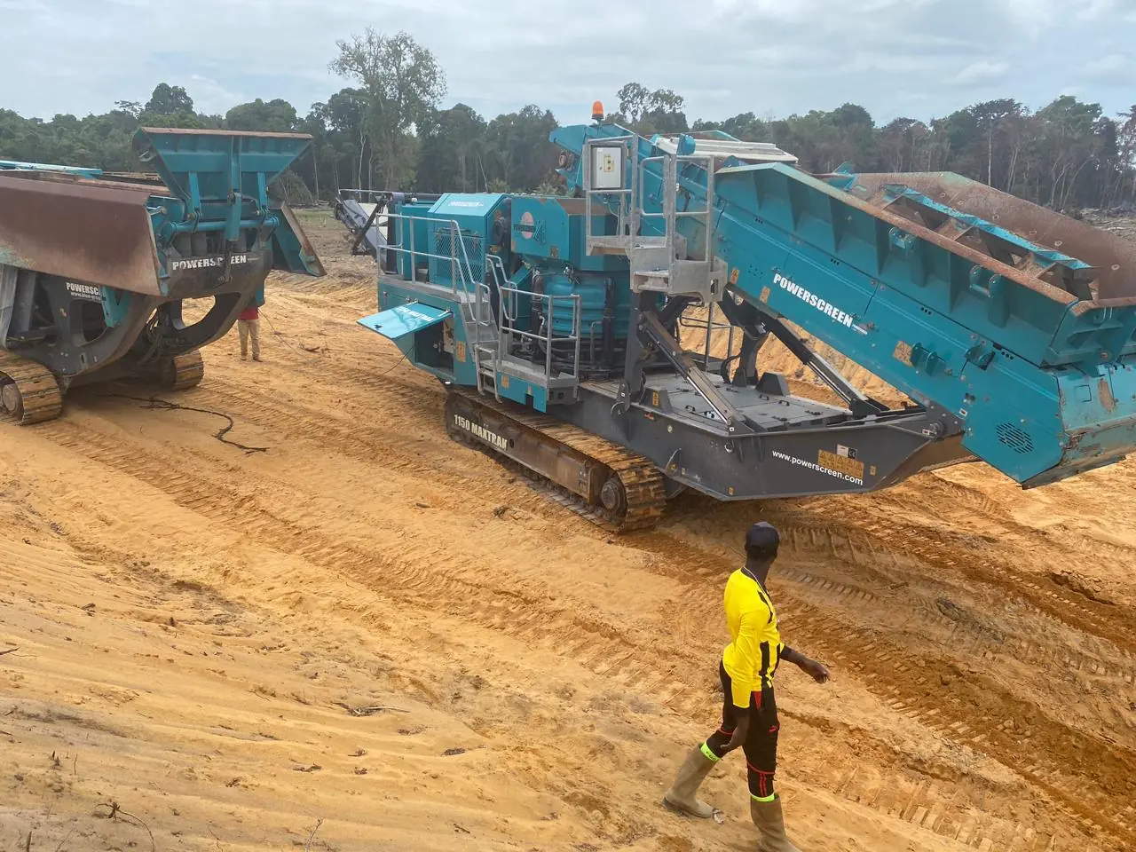 A red and grey Powerscreen 1150 Maxtrak crusher on sand, a site crew member walking past in the foreground.