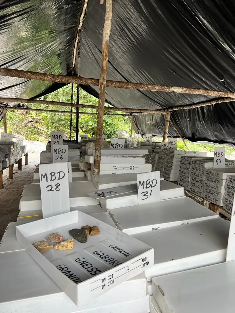 Wide view under the tarpaulin shelter — MBD25 through MBD49 core boxes stacked on pallets, with the labelled Laterite/Gabbro/Gneiss/Granite sample tray in the foreground.