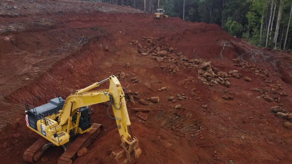 Drone view of a yellow PC490 excavator and dozer stripping red laterite overburden. Boulders break the surface on the cleared pit floor.