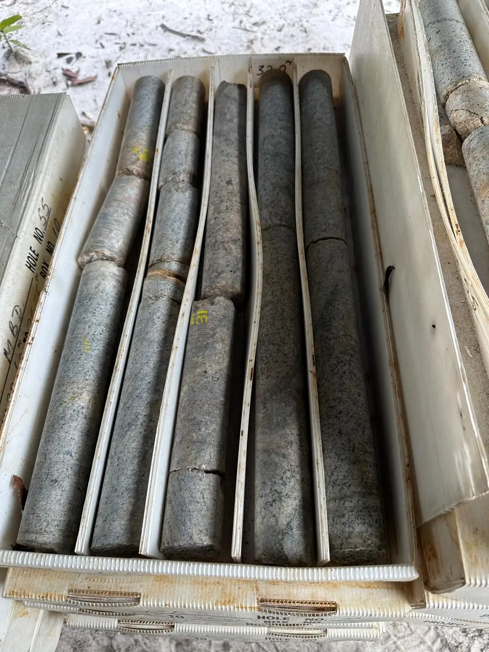 Five dark grey drill cores laid out in a numbered MBD box on white sand at the drill pad.