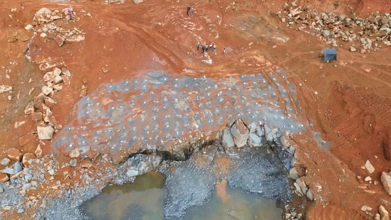 Aerial of a loaded blast pattern — a grid of black drill holes across the quarry face, crew standing back.