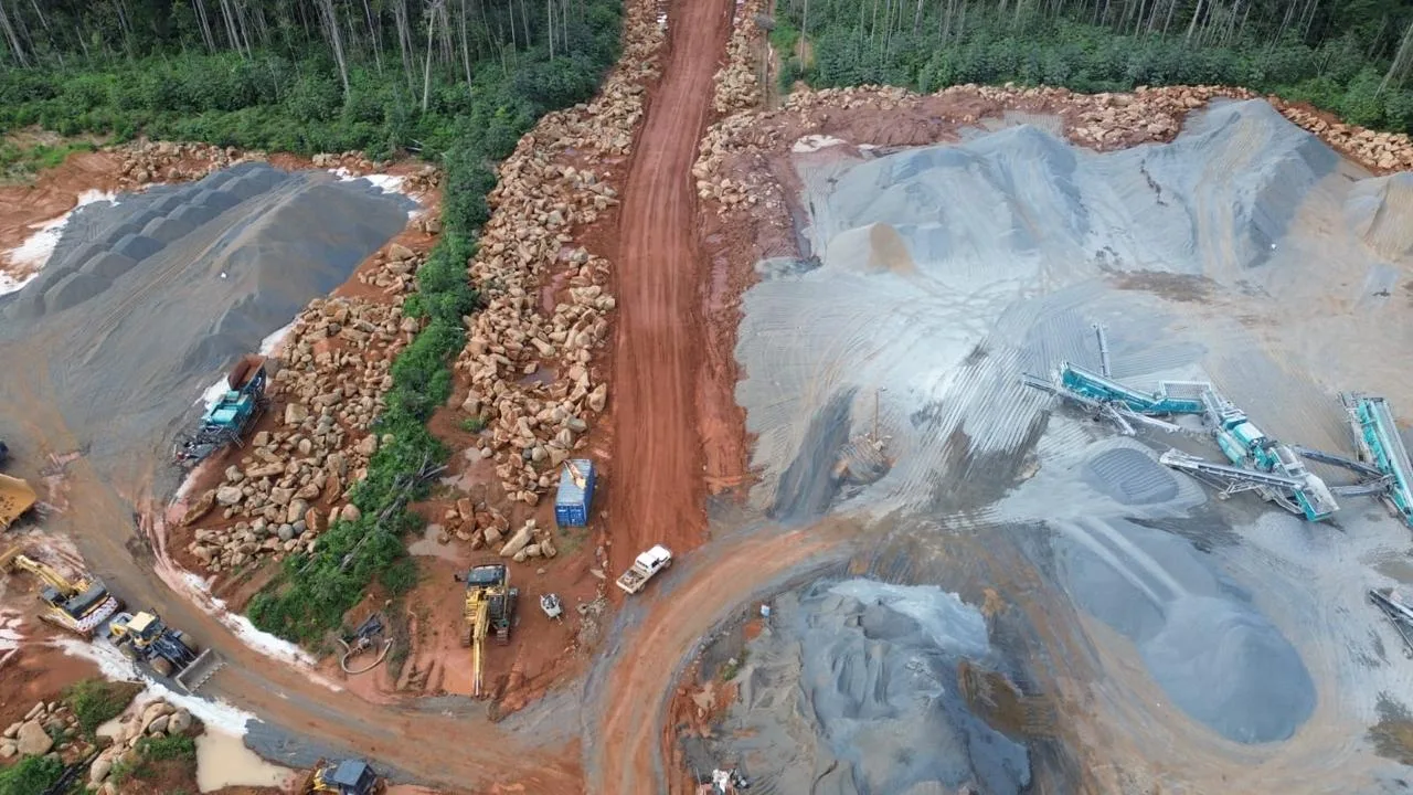 High aerial of the working quarry — crushed aggregate stockpiles at right, boulder stacks at left, the Powerscreen plant running, a red access road curving in from above.
